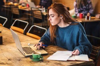 Young woman with long red hair sitting at table, working on laptop computer.